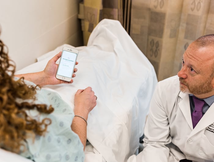 A patient in a hospital bed looking at their medical records on an iPhone while speaking to a doctor.