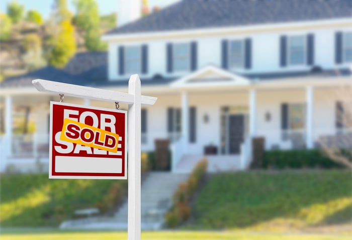 A home in the background with a "For Sale" sign with "sold" sticker pasted on it in the foreground.