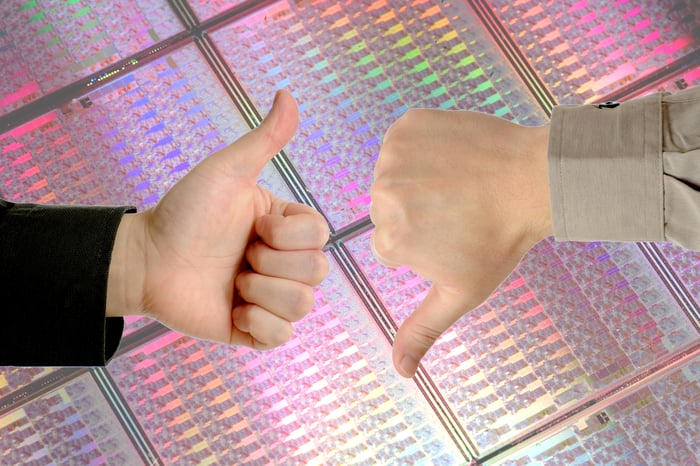 Two hands in front of an uncut wafer of silicon chips. One hand gives a thumbs-up sign and the other one signals thumbs-down.