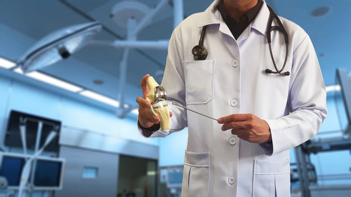 A man in a white lab coat holding a total knee replacement artificial joint.