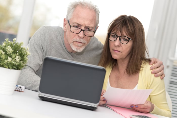 An older couple with concerned expressions sit at a table and look at a laptop screen.