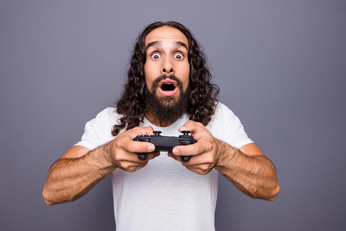 A man with long hair holds a gaming controller and is surprised facing into the camera. 