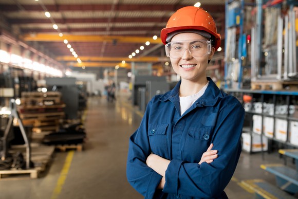 A female technician in a hard hat smiles into the camera inside a modern factory.