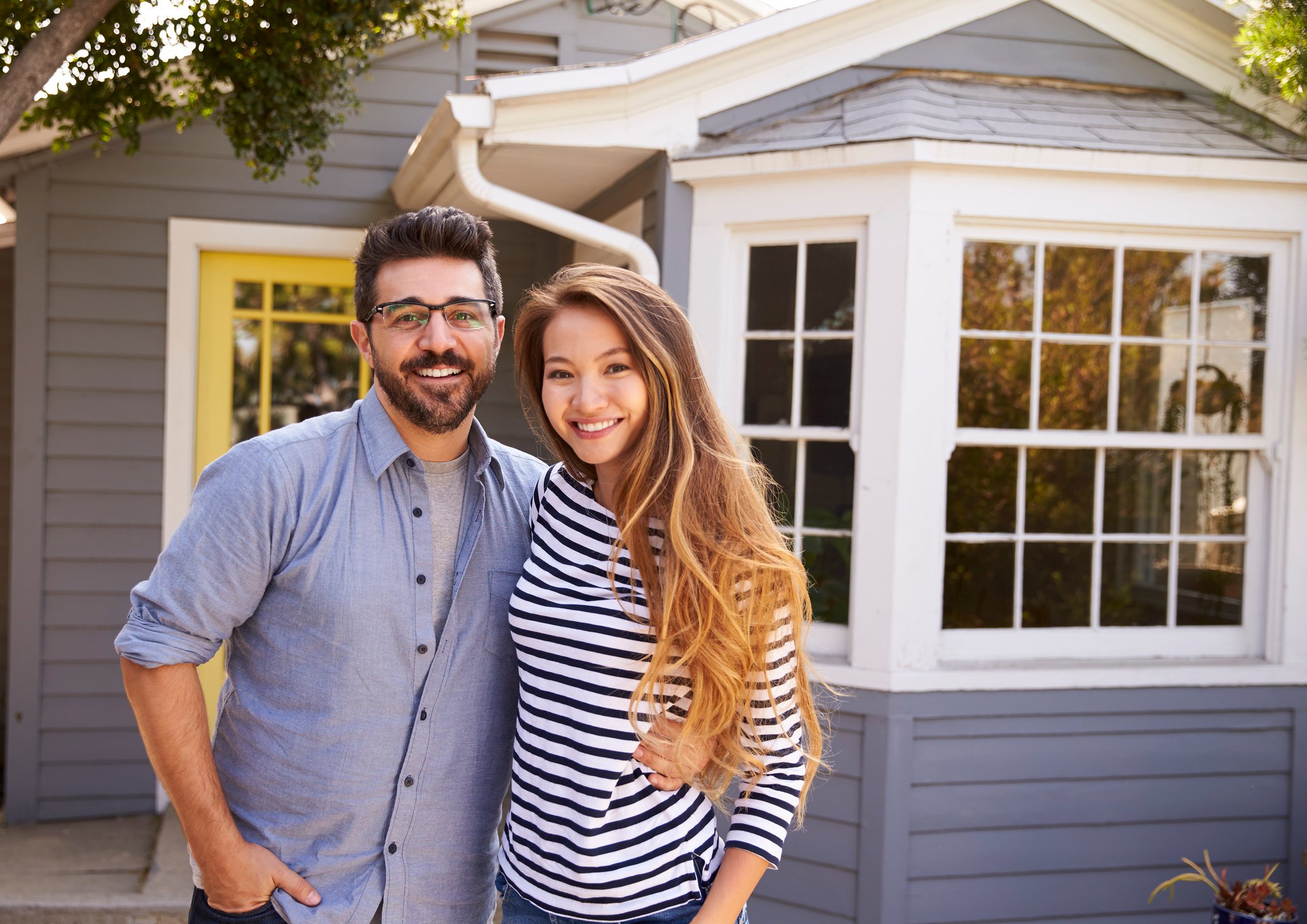 young man and woman standing in front of house with arms around each other and smiling -- couple new home homeownership real estate POC
