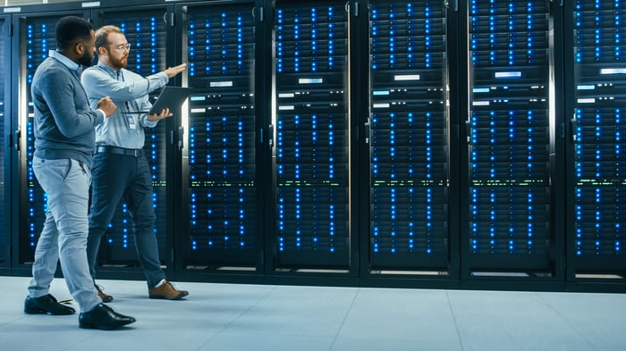 IT Technician with a Laptop Computer and Engineer Colleague are Talking in Data Center while Walking Next to Server Racks. Running Diagnostics or Doing Maintenance Work.