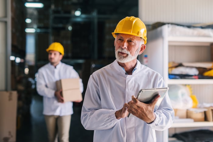 An older man in an industrial setting wears a yellow hard hat while cradling a tablet.