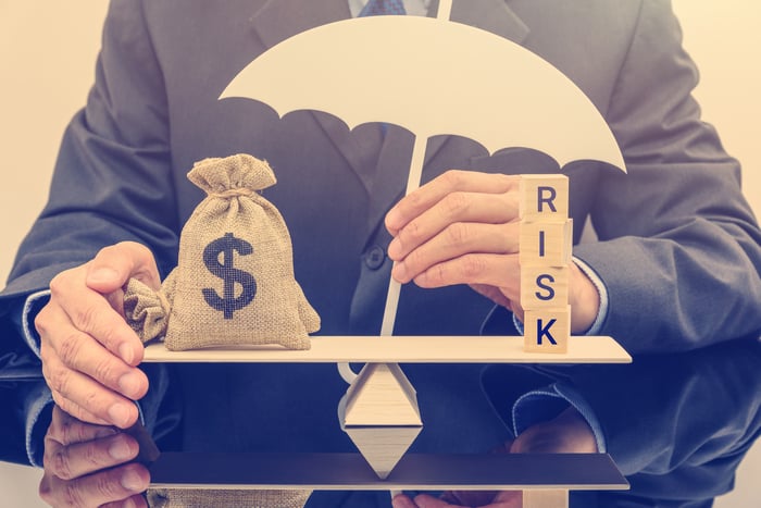 A man holding a small umbrella over a bag with a dollar sign on it and four blocks that spell out "risk" -- both balancing on a board in front of him