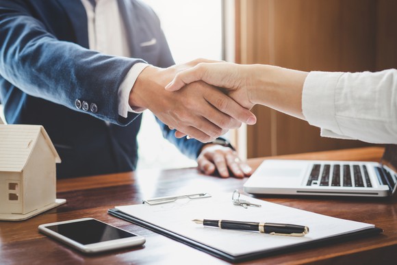 Two people shaking hands over a contract next to a mobile phone and a model of a house.
