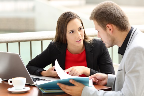 A young businessman pointing out something in a file folder he's holding. A young woman across the bistro table is asking questions.