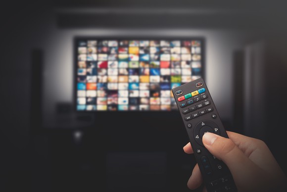 A hand holding a television remote control in the foreground with a large television on in the background