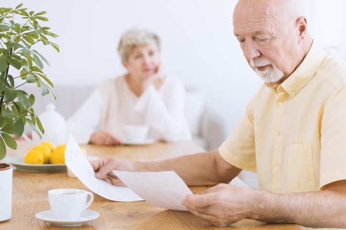 Older man looking at documents with blurry image of older woman in background