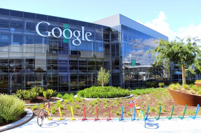 Glass-walled, multistory Googleplex with "Google" written on side of building and a couple bikes in multicolor bike racks in foreground