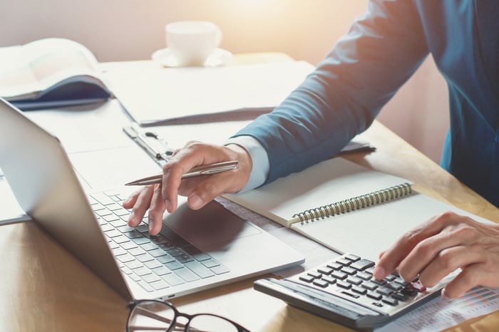 Person holding pen typing on laptop while using calculator.