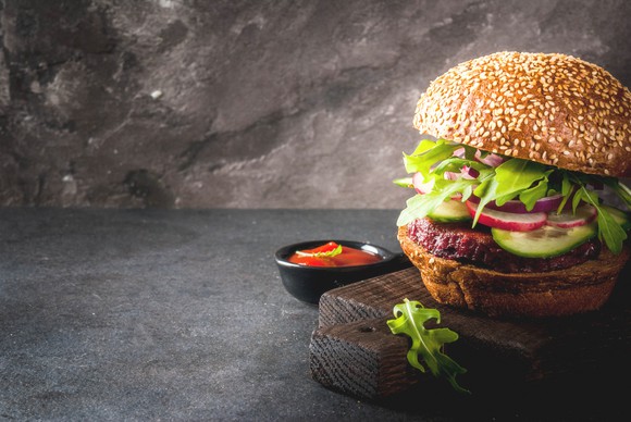 A veggie burger with lettuce and a sesame seed bun sits on a wooden surface against a dark gray background.