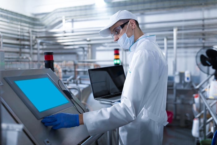 A man in a white robe and a mask with a laptop in his hands stands near the digital screen at a factory.