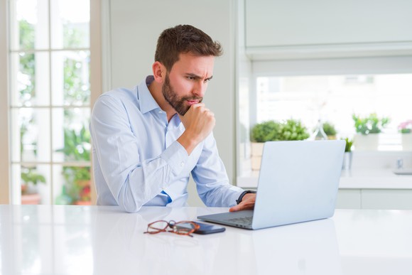 Man with serious expression resting chin on hand while looking at laptop.