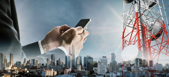 A person holding a cell phone with a city scape in the background and a cell tower.