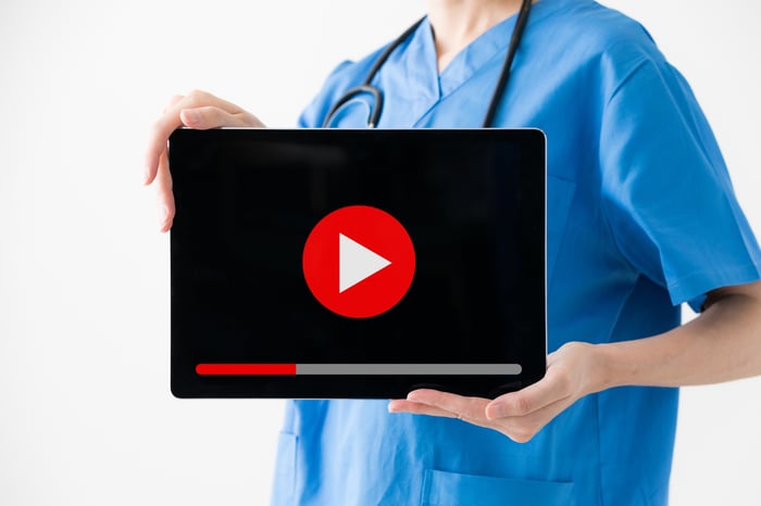 Healthcare professional in blue scrubs with a stethoscope around her neck holding a monitor with a play button displayed for a video