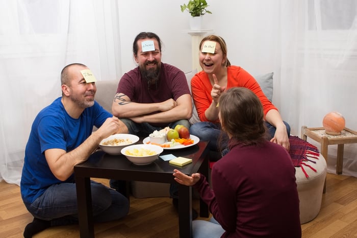 Four friends are around a table, with cards on their foreheads, playing a game and laughing.