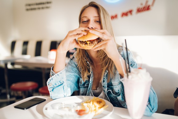 A young woman eating a burger and shake inside a restaurant.