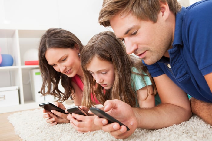 Mother, father, and daughter laying next to each other on a white rug at home with each person looking on their mobile phone.