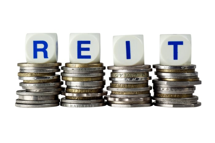 Four white cubes with the blue letters REIT, each sitting atop a pile of coins.