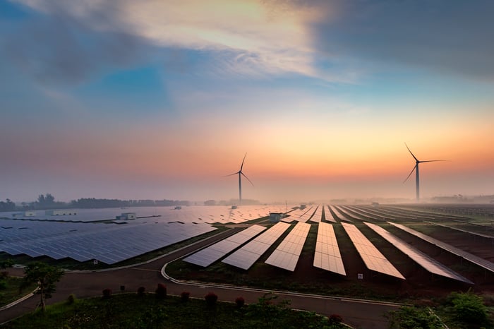 A field of solar panels with wind turbines in the background at dawn.