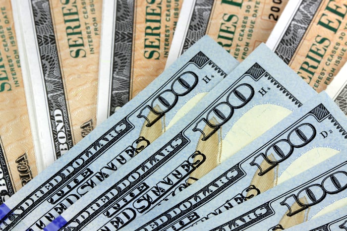 A small fanned pile of one hundred dollar bills lying atop a larger fanned pile of U.S. Treasury bonds.