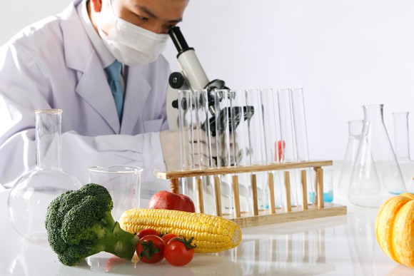 A scientist looking into a microscope with test tubes and some vegetables in the foreground.