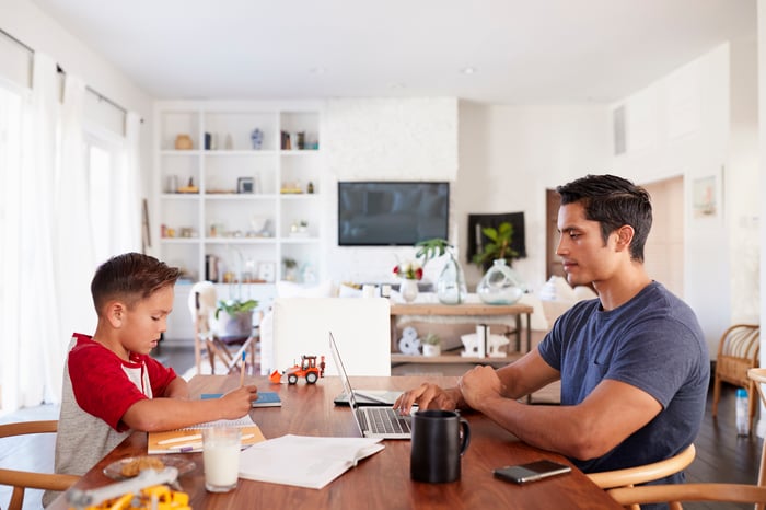 Man at table typing on laptop while young boy across from him writes in notebook