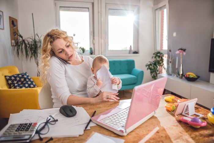 Woman at laptop balancing cell phone on ear while holding young child in lap