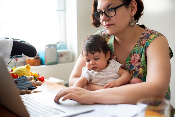 Woman with serious expression typing on laptop while holding baby