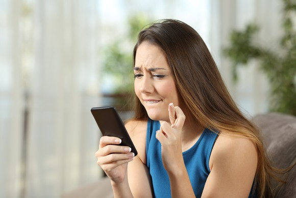 A young woman crosses her fingers and bites her lip while frowning at her smartphone.