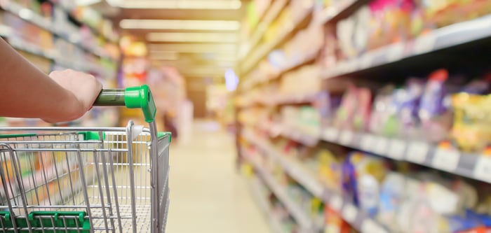 A shopper pushing a grocery cart in a supermarket aisle with products on shelves blurred.