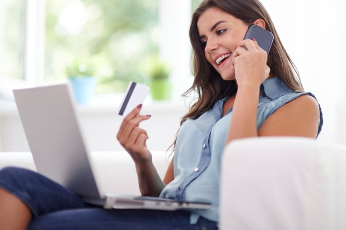 A woman talking on her smartphone and holding a credit card with an open laptop of her lap.