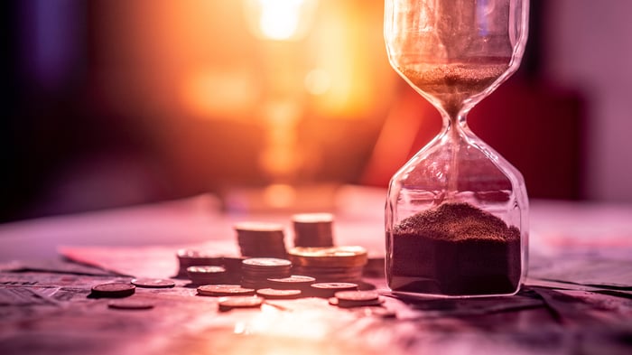 Sand running through an hourglass, which is surrounded by stacks of coins and messy cash bills laid across a table. 