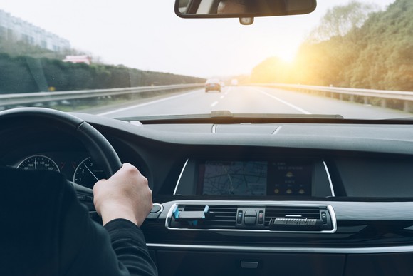 View of car dashboard, hand on wheel, and road ahead.
