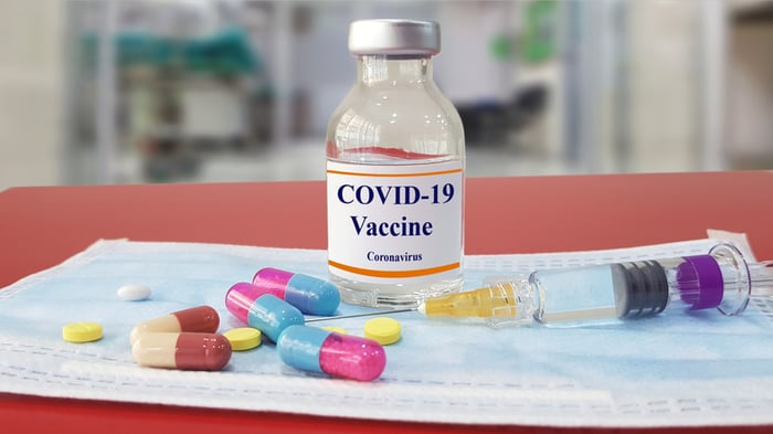 Vial labeled 'COVID-19 vaccine,' a syringe, and a few pills sitting on a flat surface.