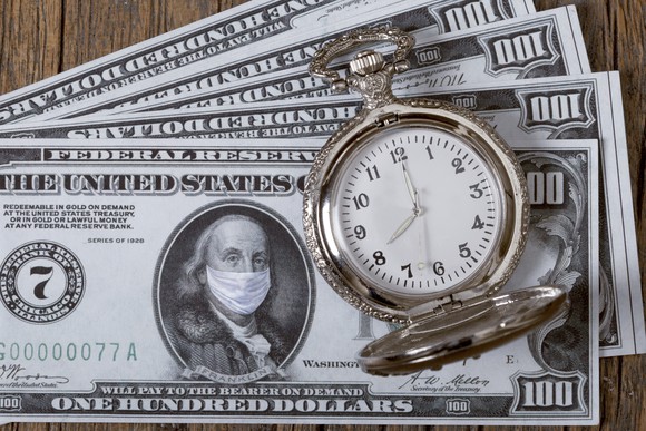 A stopwatch sits atop a pile of hundred dollar bills, featuring Ben Franklin wearing a medical mask.