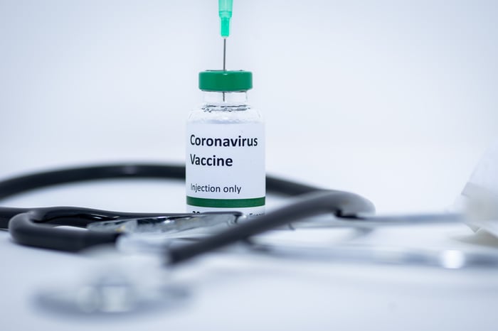 Small vial with "Coronavirus vaccine" written on it and a stethoscope sitting on a flat white surface.