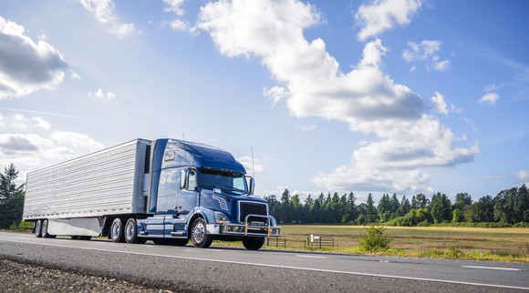 A refrigerated semi truck driving along a tree-lined highway.