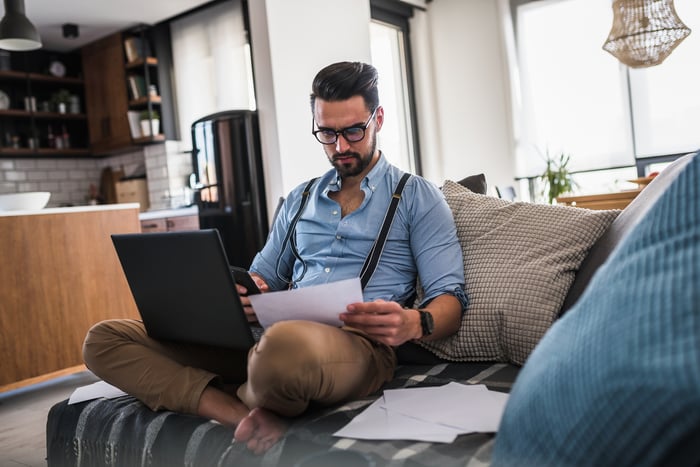 Man sitting on couch with laptop in lap holding phone and document