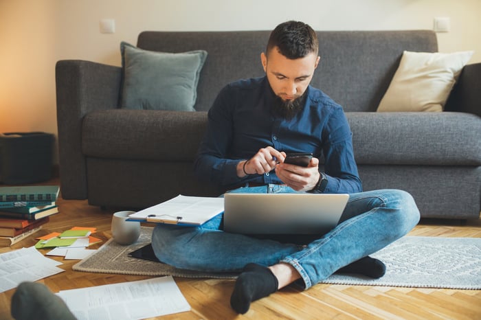 Man sitting on floor holding phone with laptop and clipboard in lap