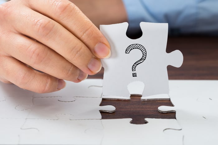 A person holding up a large white puzzle piece with a question mark drawn on it.