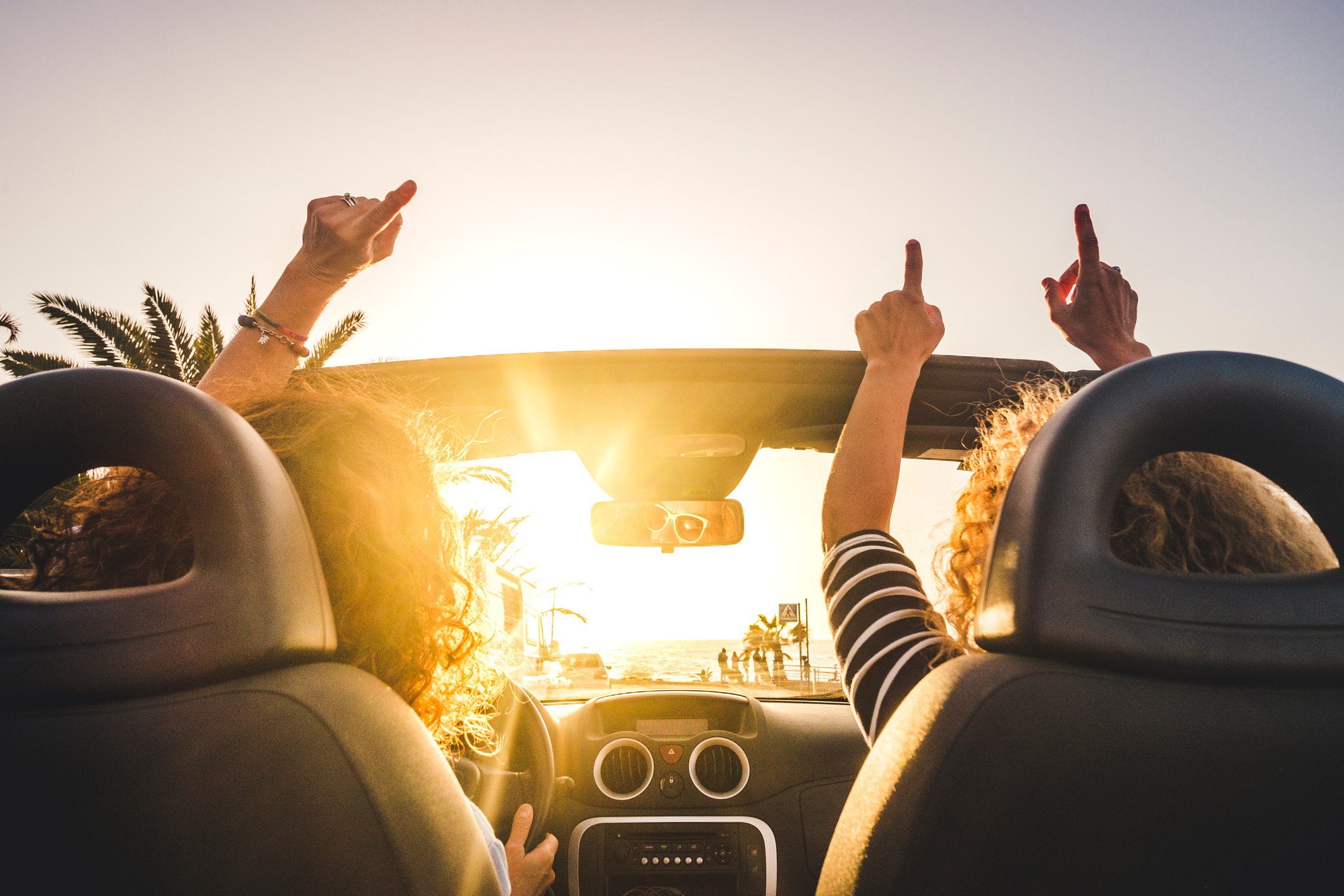 Two women driving in a convertible with their hands in the air listening to music