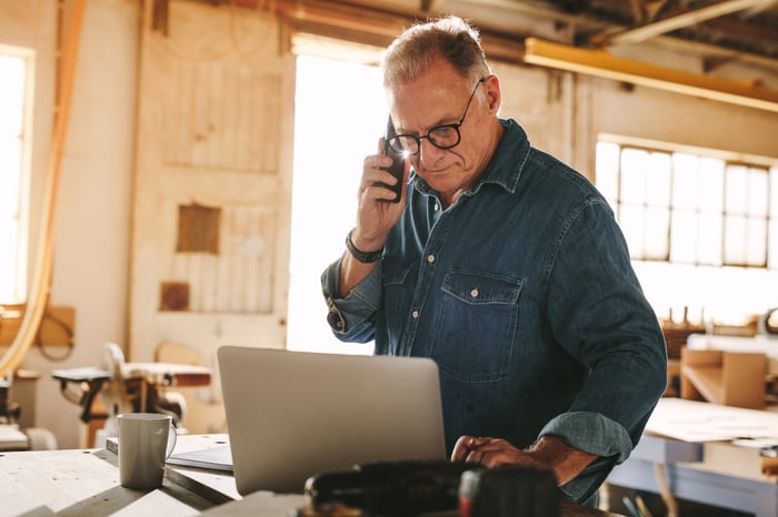 Older man standing at laptop holding phone to ear