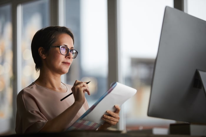 A mature woman writes on a notepad in front of her desktop computer monitor in an office. 