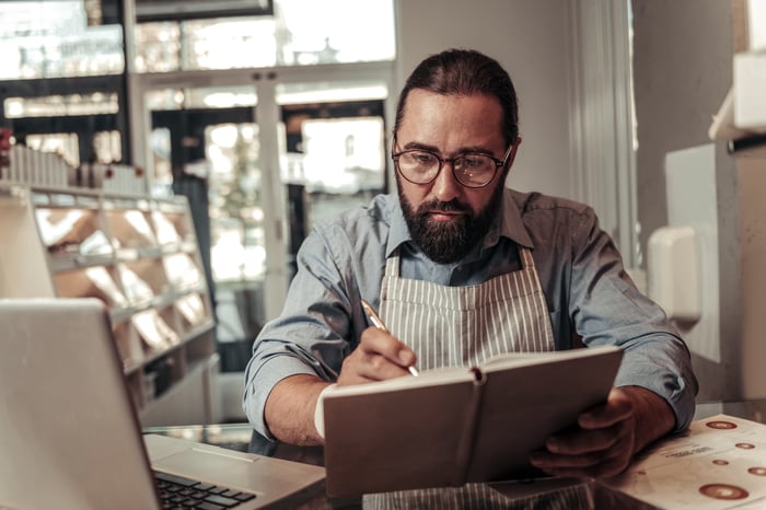 Man in apron with serious expression writing in notebook next to laptop