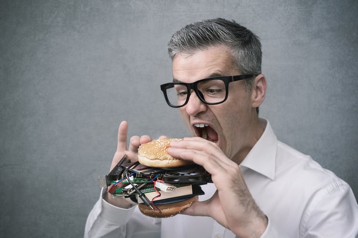 A tech nerd opens his mouth to eat a hamburger made of computer parts.