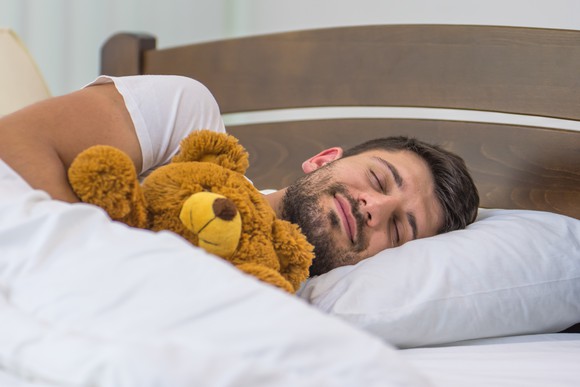 A grown man sleeps soundly in bed, squeezing a stuffed teddy bear.